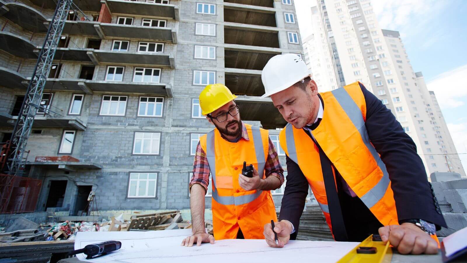 Two construction workers in hard hats and safety vests review blueprints on a table at a construction site. The unfinished building towers behind them.