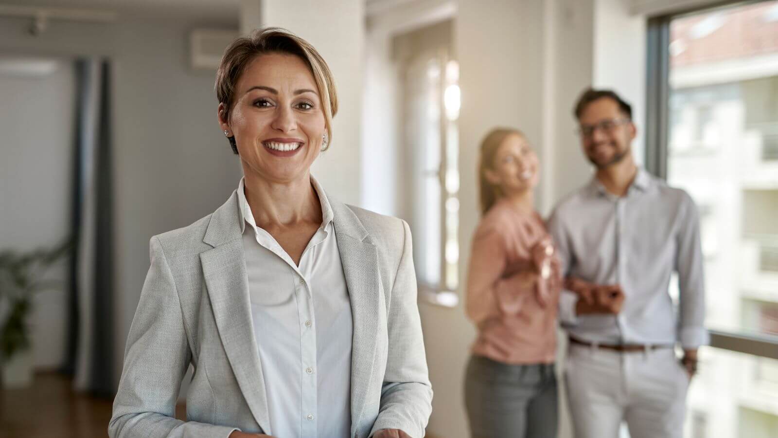 A woman in a light gray suit jacket and white shirt smiles at the camera, with a blurred couple standing behind her near a window.