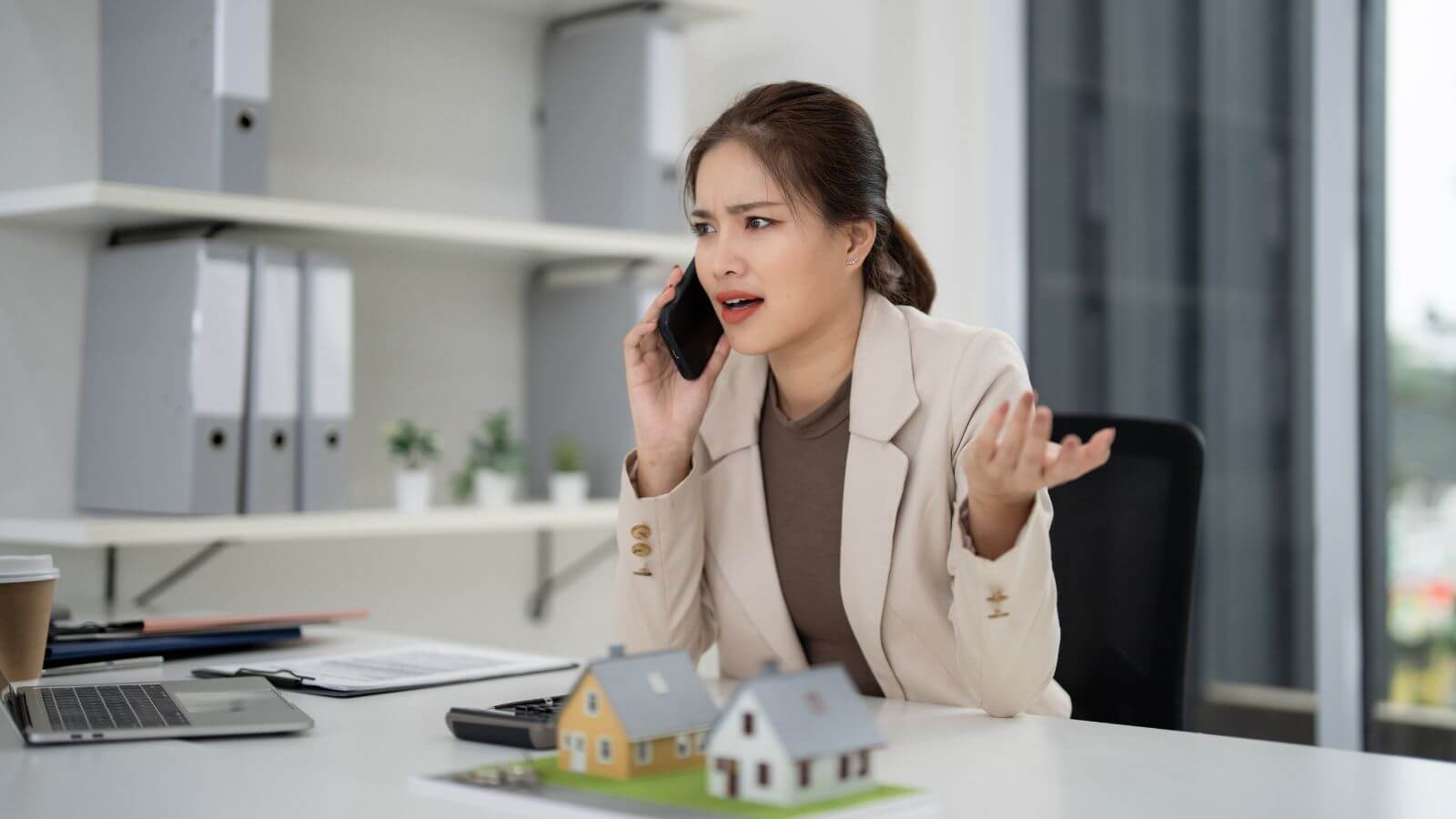 A person in a beige blazer is talking on a mobile phone while gesturing with their other hand, sitting at a desk with miniature houses, a laptop, and a coffee cup.