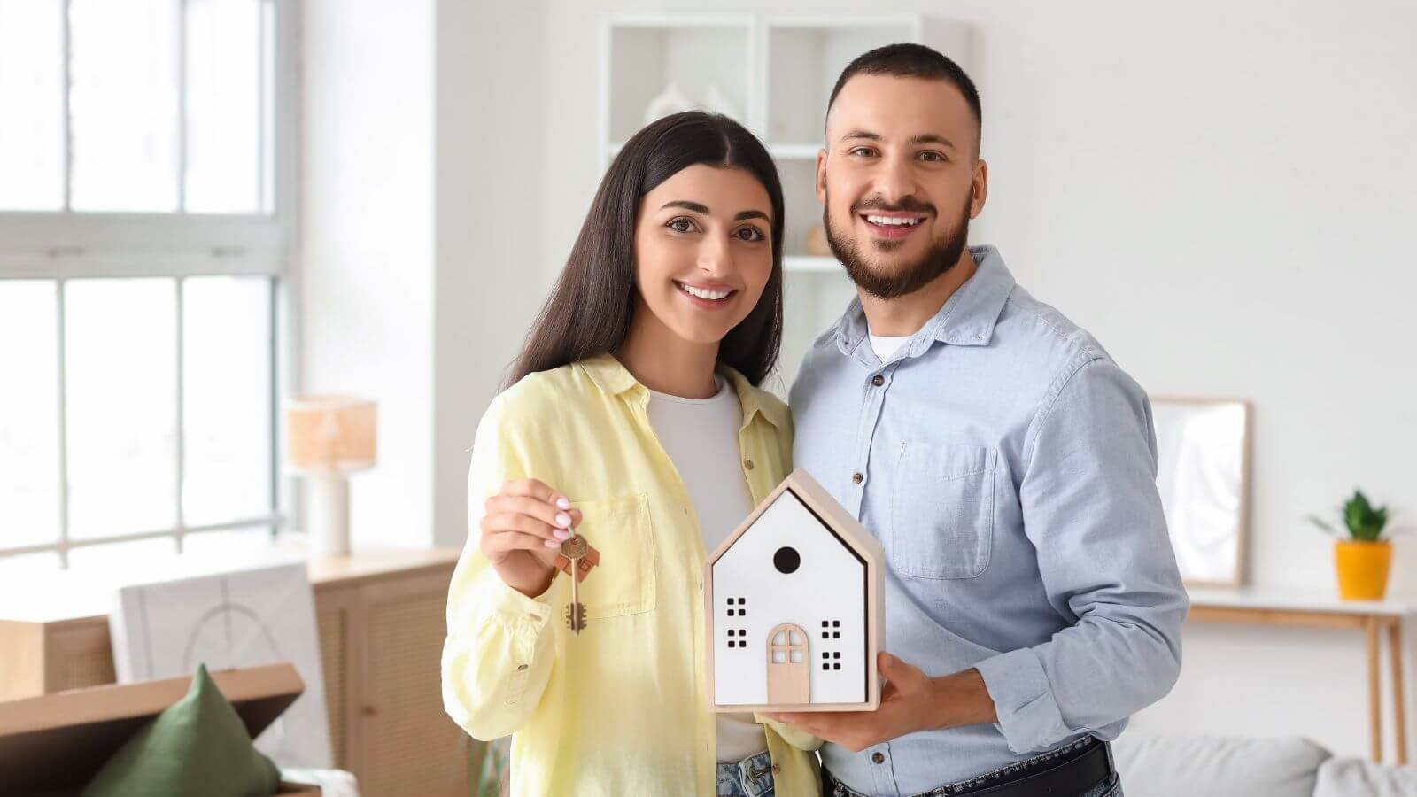 A smiling couple holds a model house and keys, celebrating buying a new home. The woman wears a yellow shirt, and the man wears a blue shirt.