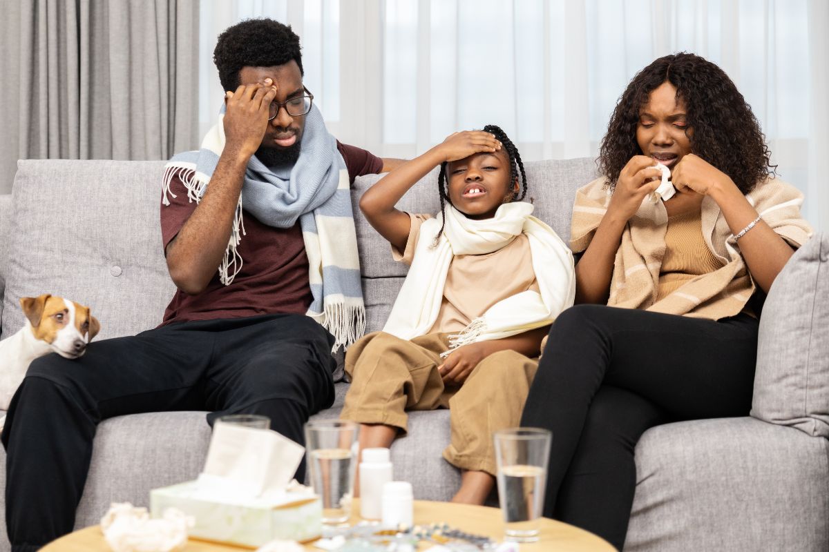 Family of three sitting on a couch with symptoms of illness, such as headaches and sneezing, indicating potential indoor air quality issues and their impact on health.