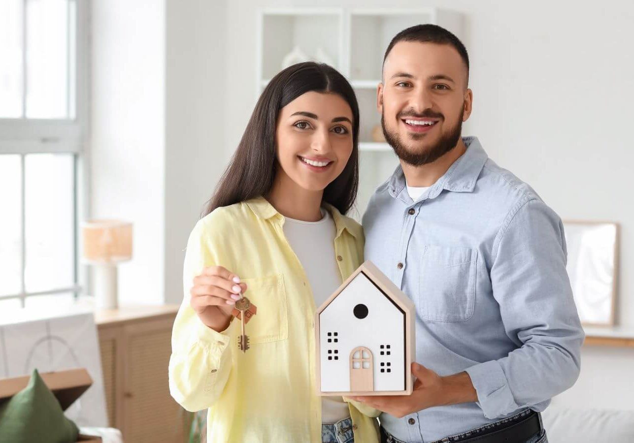 A smiling couple holds a model house and keys, celebrating buying a new home. The woman wears a yellow shirt, and the man wears a blue shirt.
