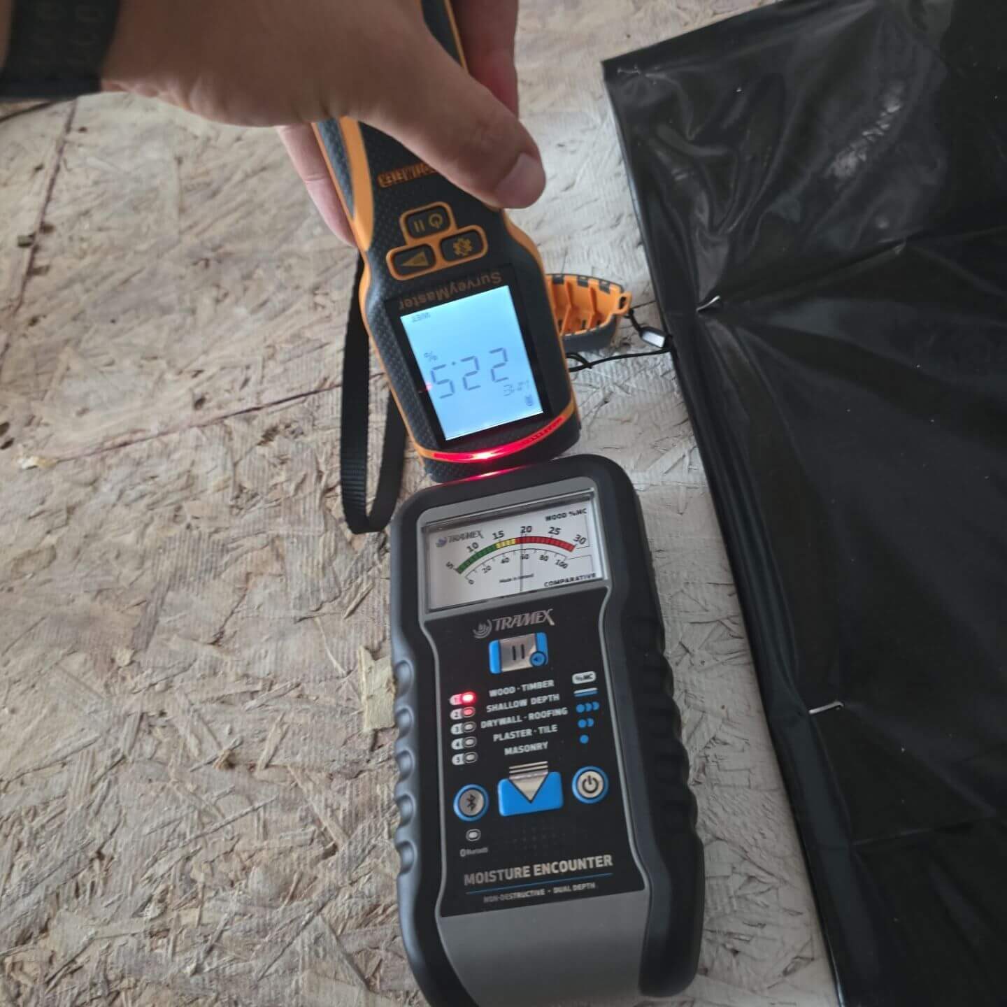 Side-by-side moisture testing using a Tramex Moisture Encounter and a pin meter on a subfloor to confirm drying progress.